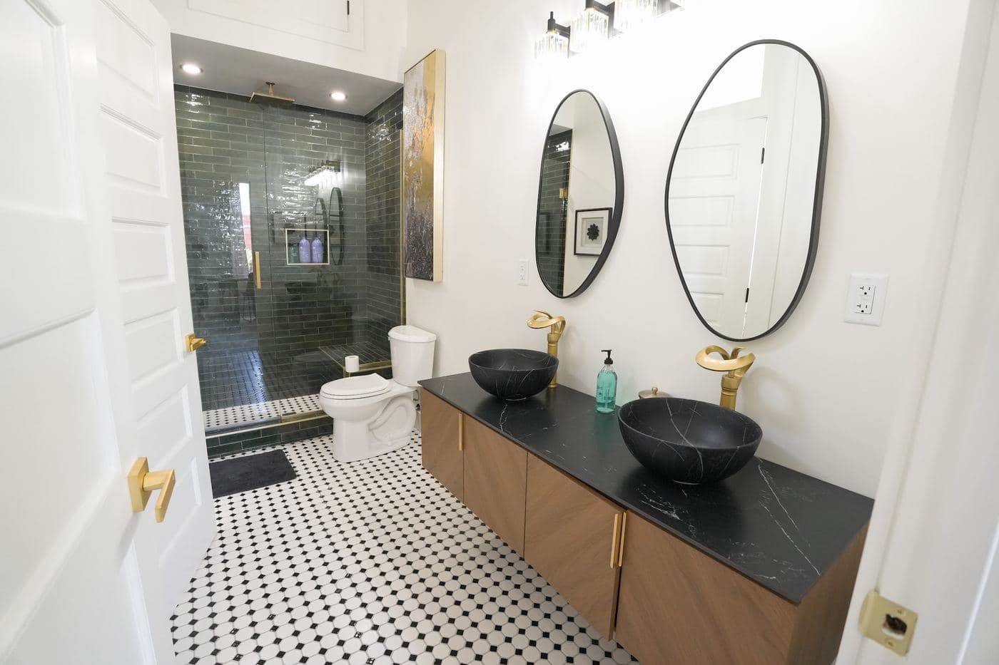 A loft bathroom with black marble vessel sinks, brass fixtures, a walnut vanity and a hunter-green tiled shower